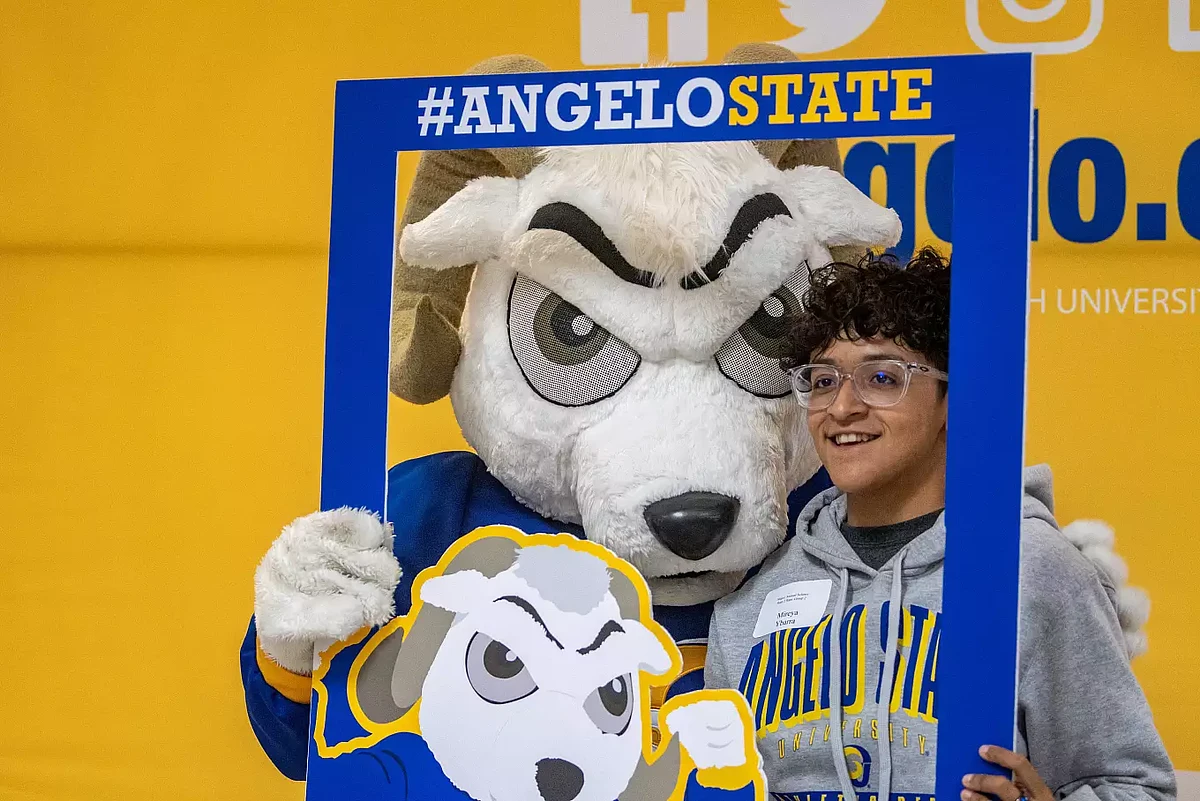 A person poses with a mascot behind a blue frame featuring "#ANGELOSTATE" against a yellow background.