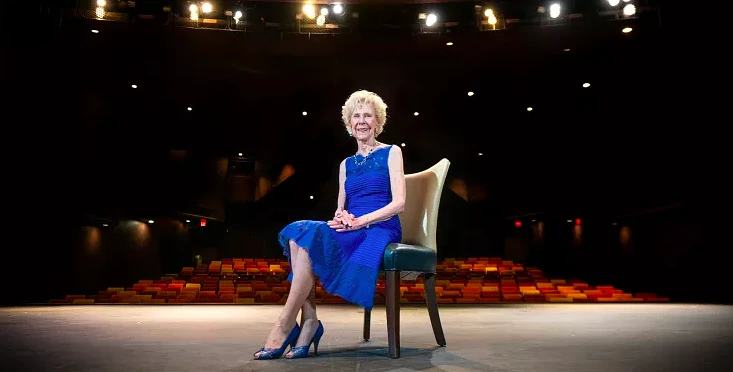 A woman in a blue dress sits on a chair in the middle of a stage