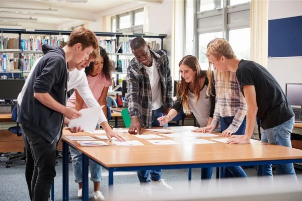 A group of young people are collaborating on a project at a table in a library or classroom setting.